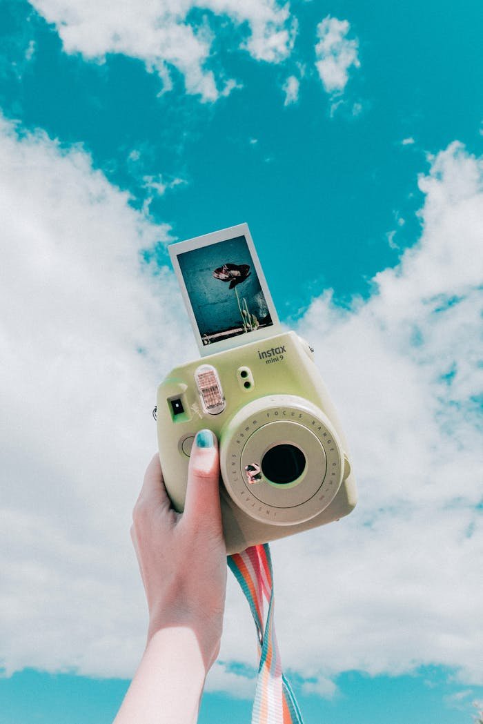 Hand holding an instant camera with a sky background, capturing a fun and retro vibe.